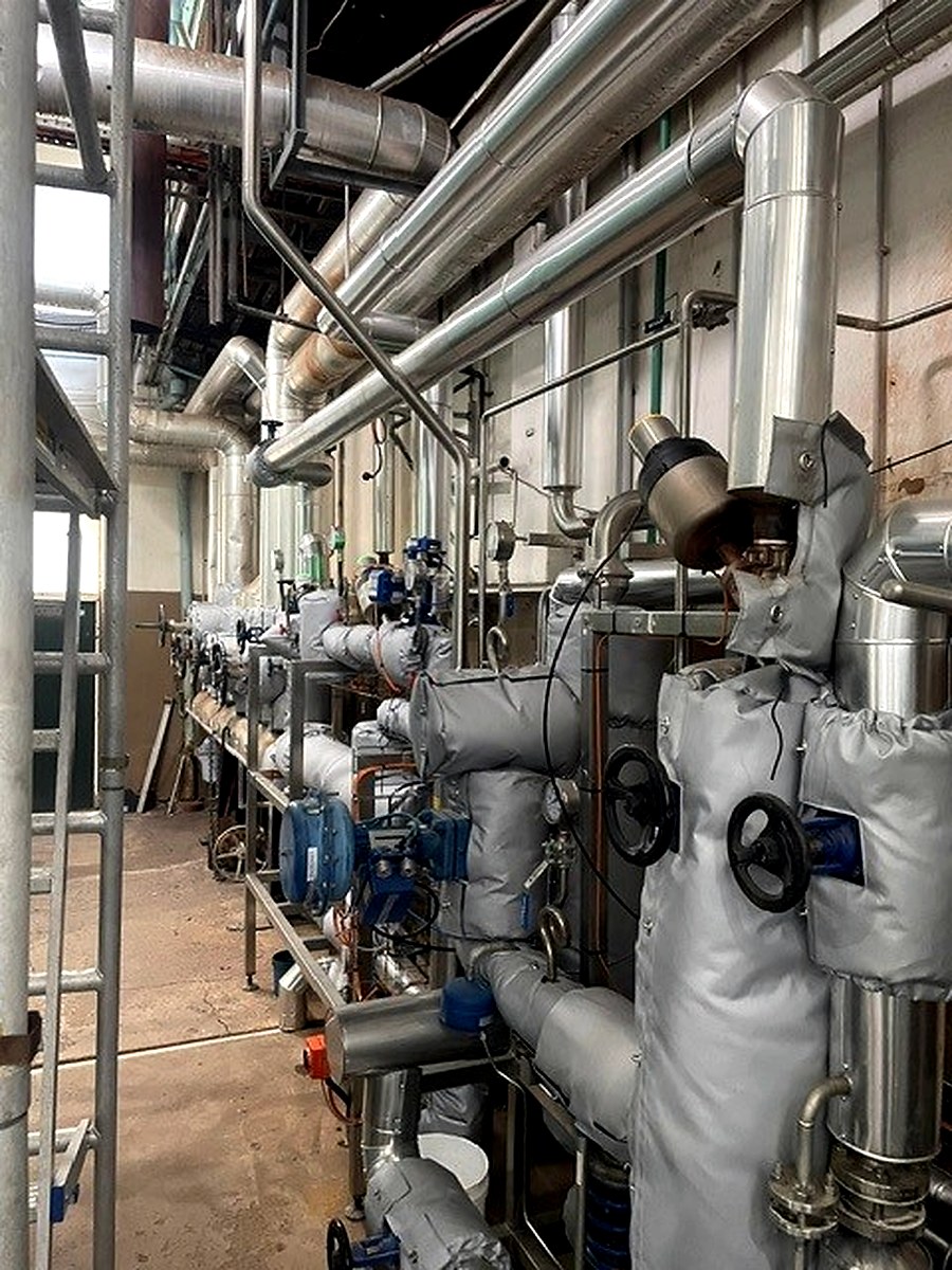 Long industrial corridor showing thermal jackets on valve assemblies along an extended pipe run